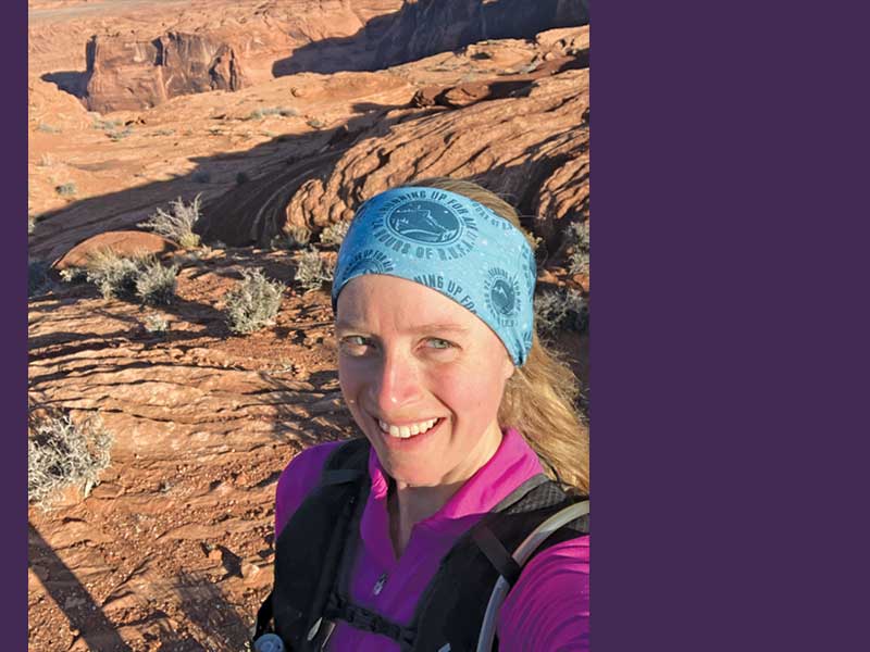 A young woman wearing a pink top and backpack, with a patterned light blue bandana standing in a desert setting with brown rocks and small plants behind her