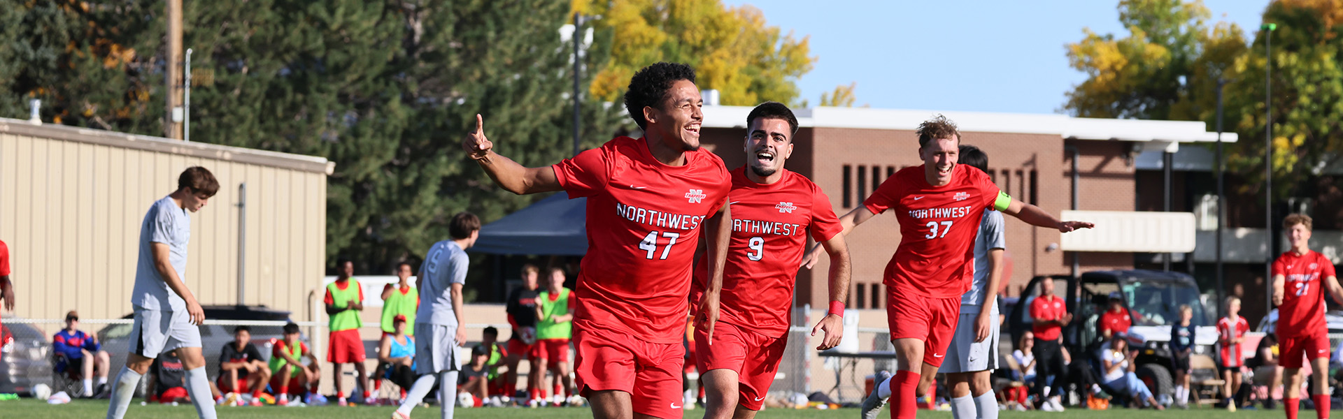 Three male soccer players running across the field celebrating a goal with buildings, spectators, and fall colors in the background