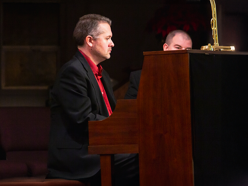A man in a black coat and redshirt playing a church piano lighted by a lamp on top