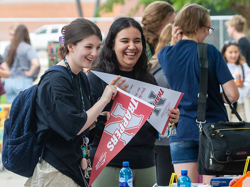 Two smiling female college students standing at a table outside holding Northwest College pennants