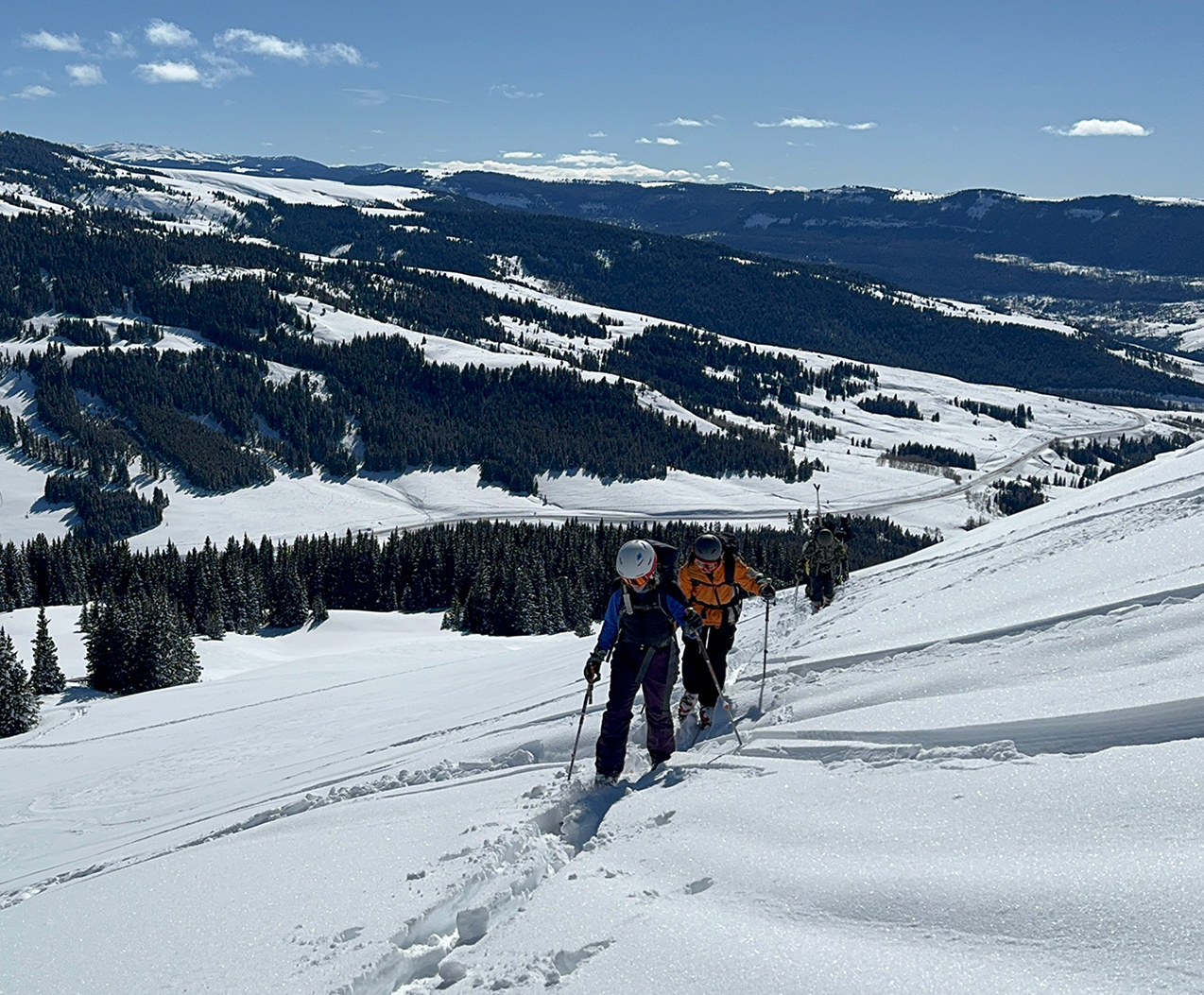 Students in ski gear moving across a snow-covered mountain with ski slopes on a mountain behind them