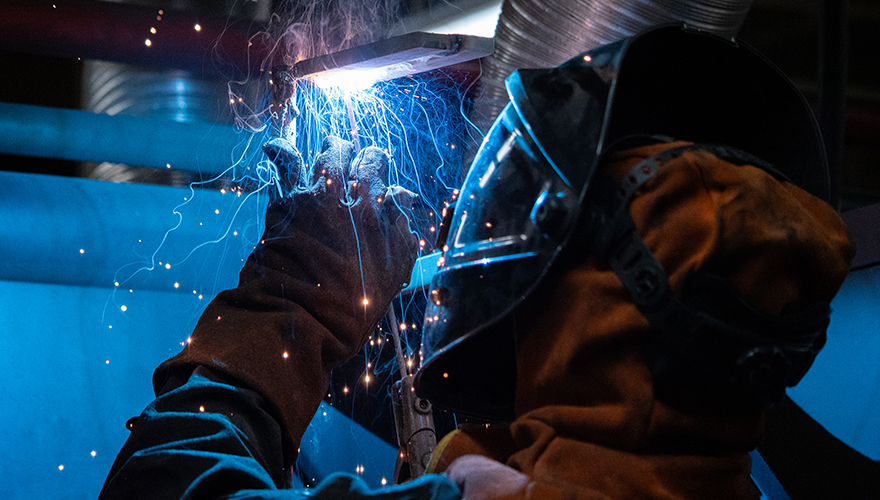 Close up shot of a standing welder in a protective hood looking upward while using a welding tool with blue sparks