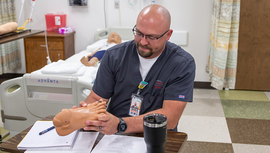 A man with a shaved head wearing glasses and a grey nursing shirt holding a replica of a foot while sitting at a desk with notebooks and a mug, all in a nursing classroom