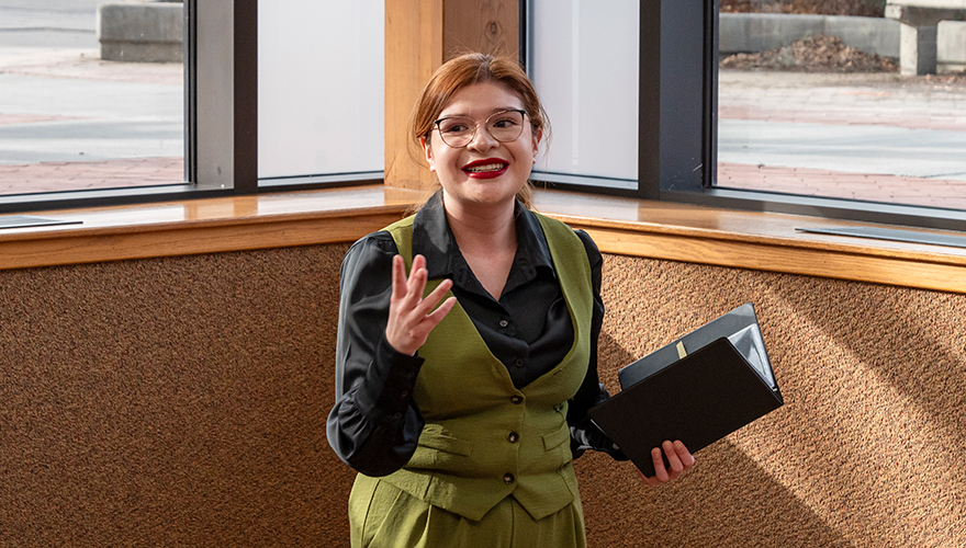 A young woman in a dark green and black suit holding a folder, and smiling while speaking and gesturing to an audience 