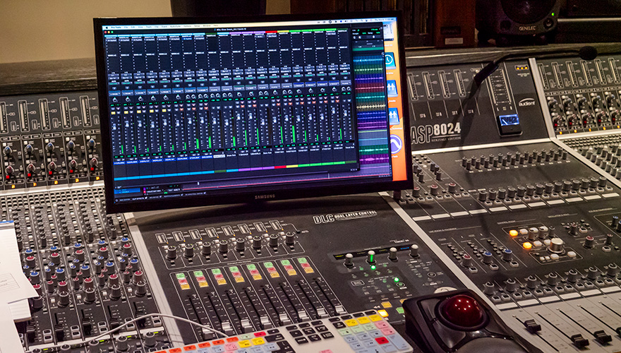 A monitor showing the digital audio controls sitting on top of a control board inside a studio
