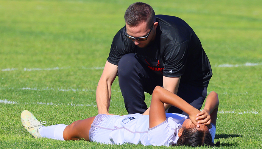 A male athletic trainer dressed in all black kneeling in the grass and checking the knee of a female soccer player laying on her back