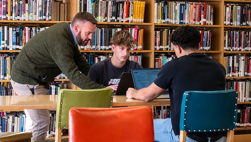 Two male students sitting a table inside a library with a male professor standing next to them looking at their laptops