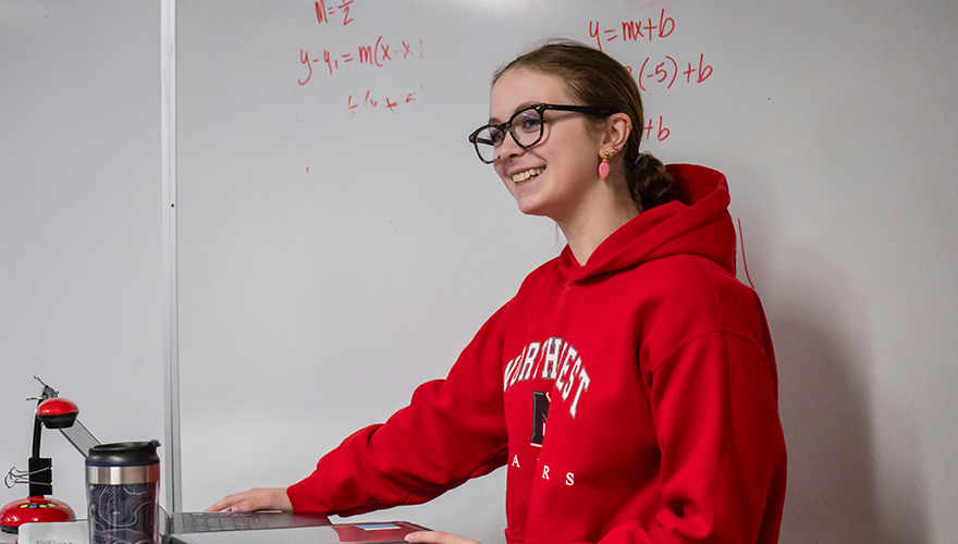 Female college student wearing glasses and a red Northwest College sweatshirt standing in front of a whiteboard with math equations talking to a class