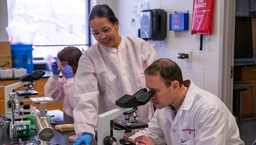 Male student sitting looking through a microscope with female student standing next to him