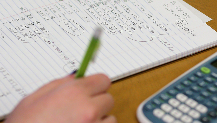 Hand holding a pen with note paper filled with numbers on a table and a calculator on the side