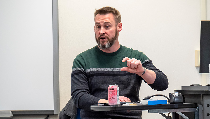 Male business professor with a beard speaking to a class while sitting behind a desk