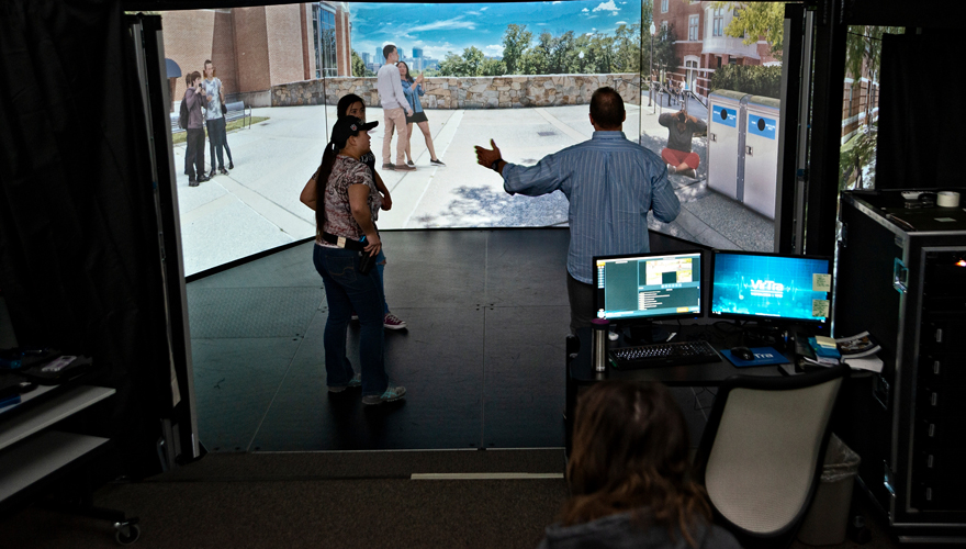 Male instructor talking to two students in front of a 270 degree simulator screen with computer control system and chair in foreground