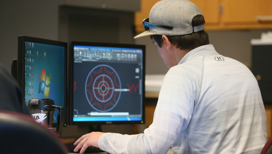 Male student with his back to the camera wearing a baseball cap sitting at two screens looking at drafting software