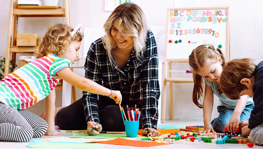 Female teacher sitting on the floor with preschool children who are coloring and playing with toys