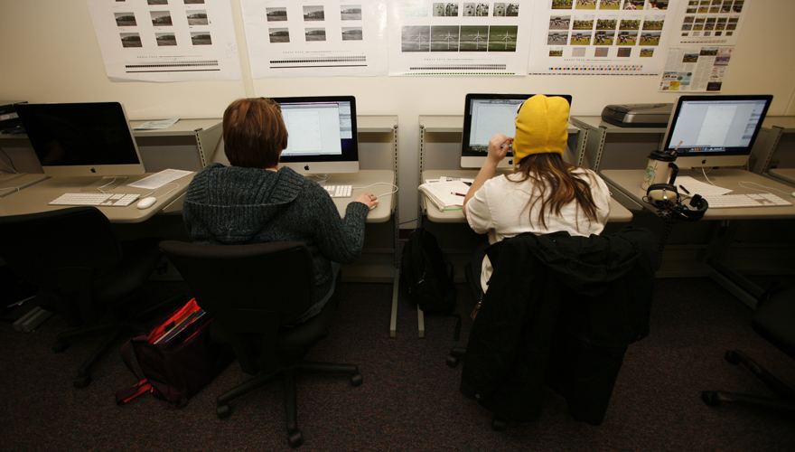 Students sitting facing computer screens in a classroom with images on the wall above them