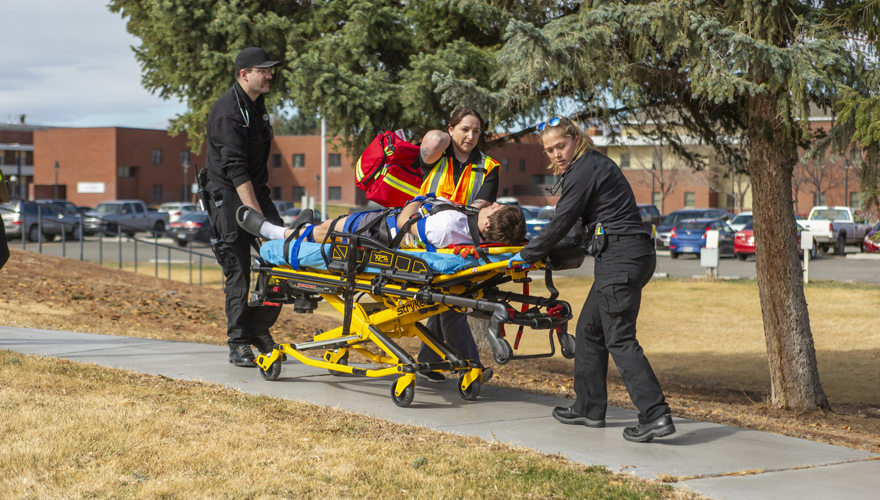 Male and female EMS students guiding a stretcher with a patient on it down a sidewalk with instructor walking beside them and trees in the background