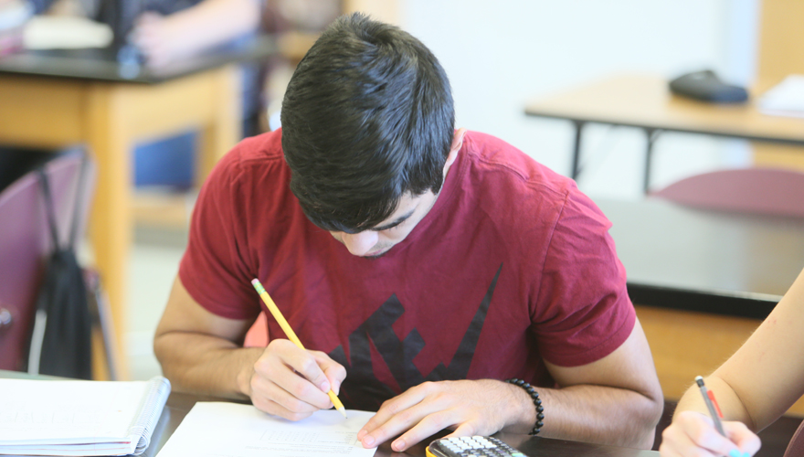 Male student at a desk writing on paper with another hand on a calculator