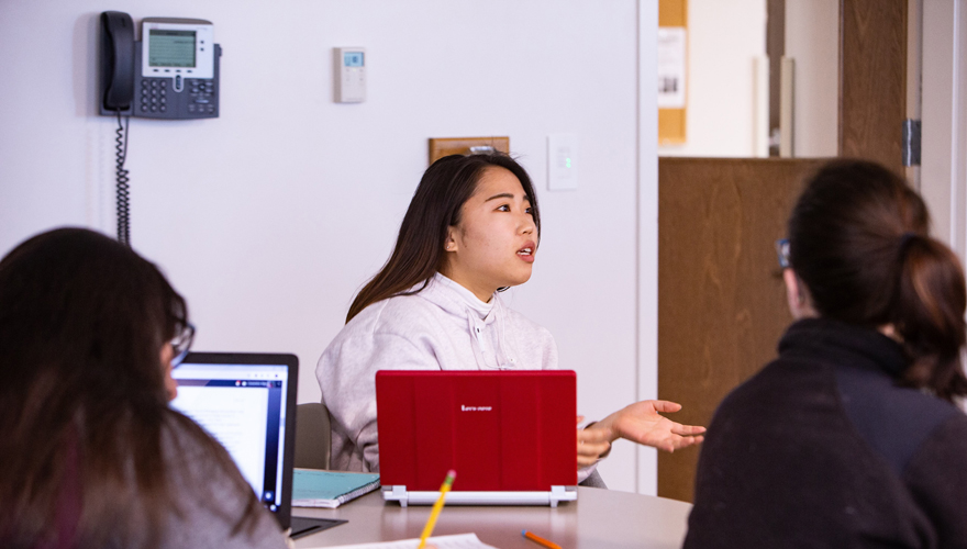 Female student sitting at a table eith two other students next to a laptop with hands out and talking to an instructor