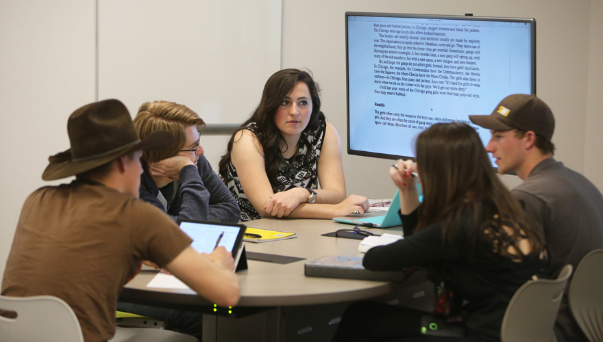 Students sitting around a table talking with a wall monitor behind them showing text