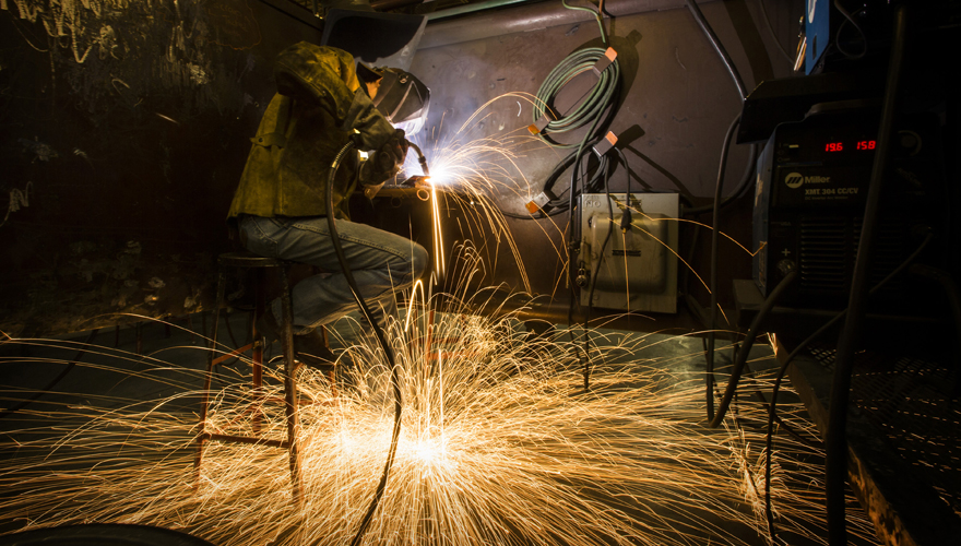 Welder sitting on a stool working on a project with bright yellow sparks showering the floor and hoses on the wall behind him