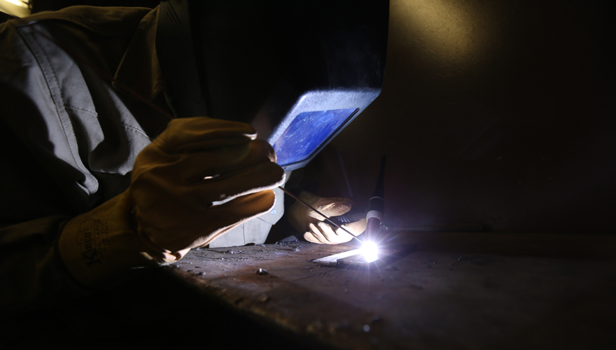 Welder wearing a heavy face shield leaning over a table doing detailed work with a brightly lit piece of metal