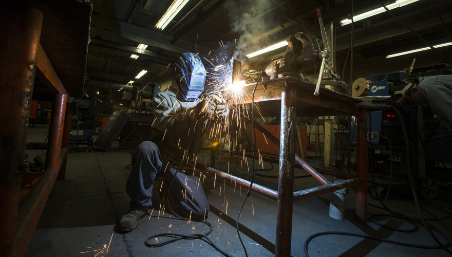 Welder in a custom designed head gear kneeling on the floor working on a project with sparks flying off a table next to him