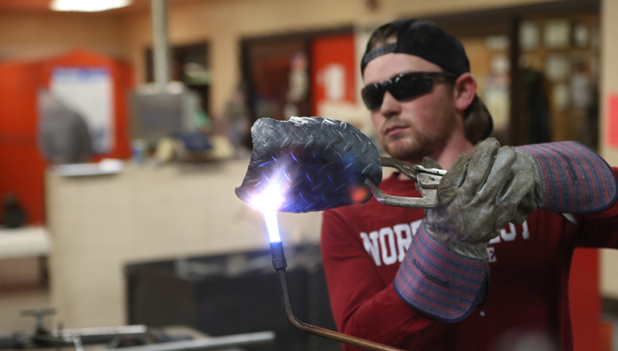 Male student in red Northwest College shirt inside a shop holding a piece of metal above a welding flame