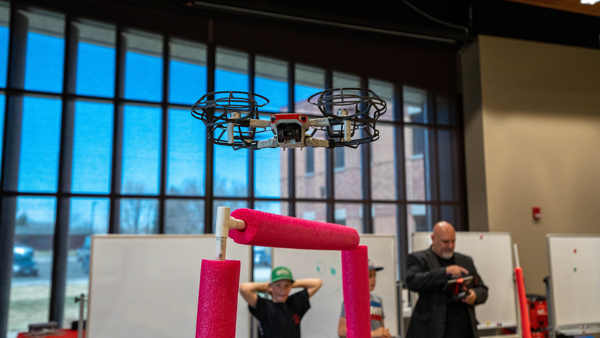 A drone hovering inside a large room with an obstacle course and people watching in the background