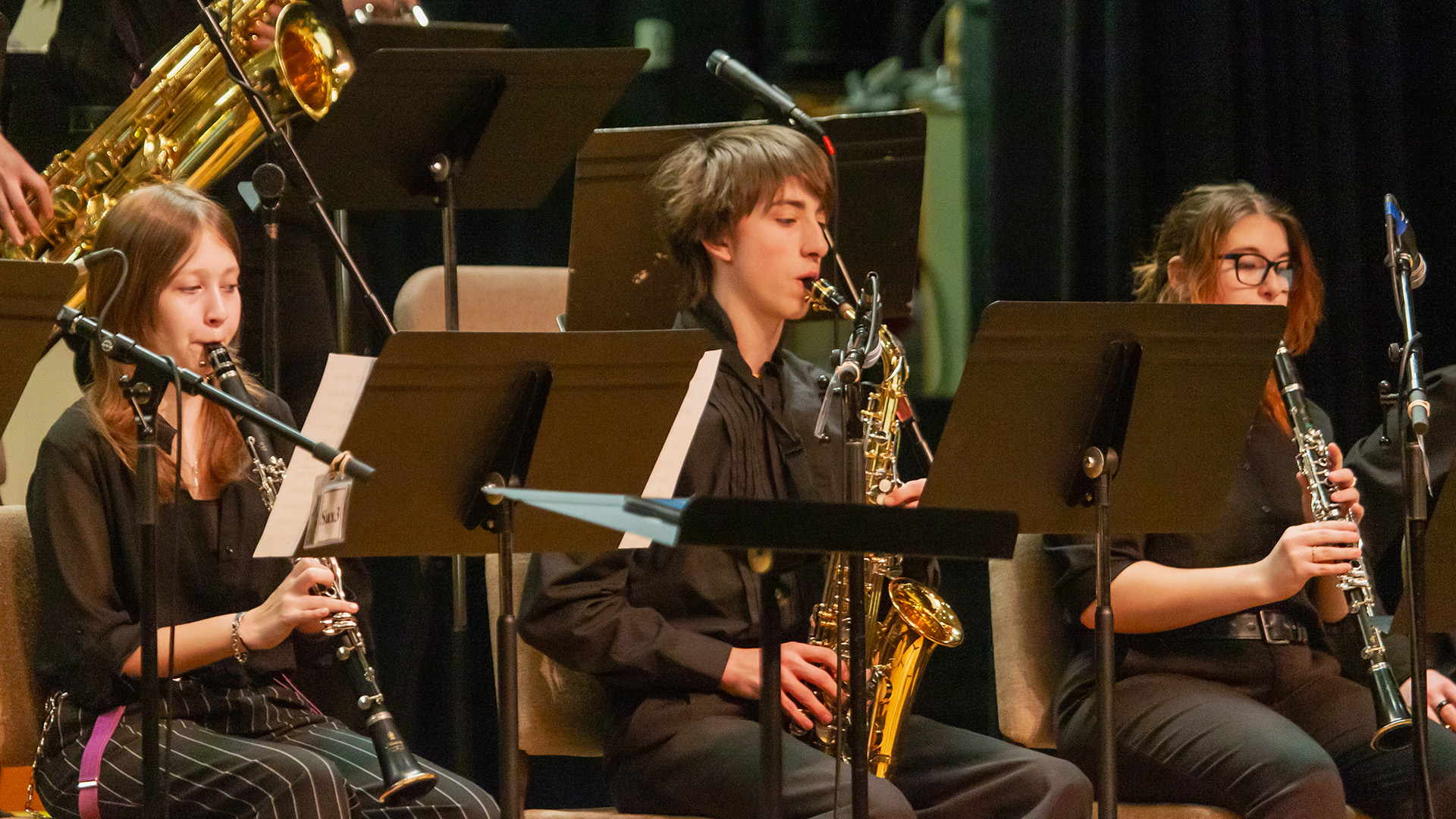 Male student playing a saxophone sitting on stage between two female students playing clarinets