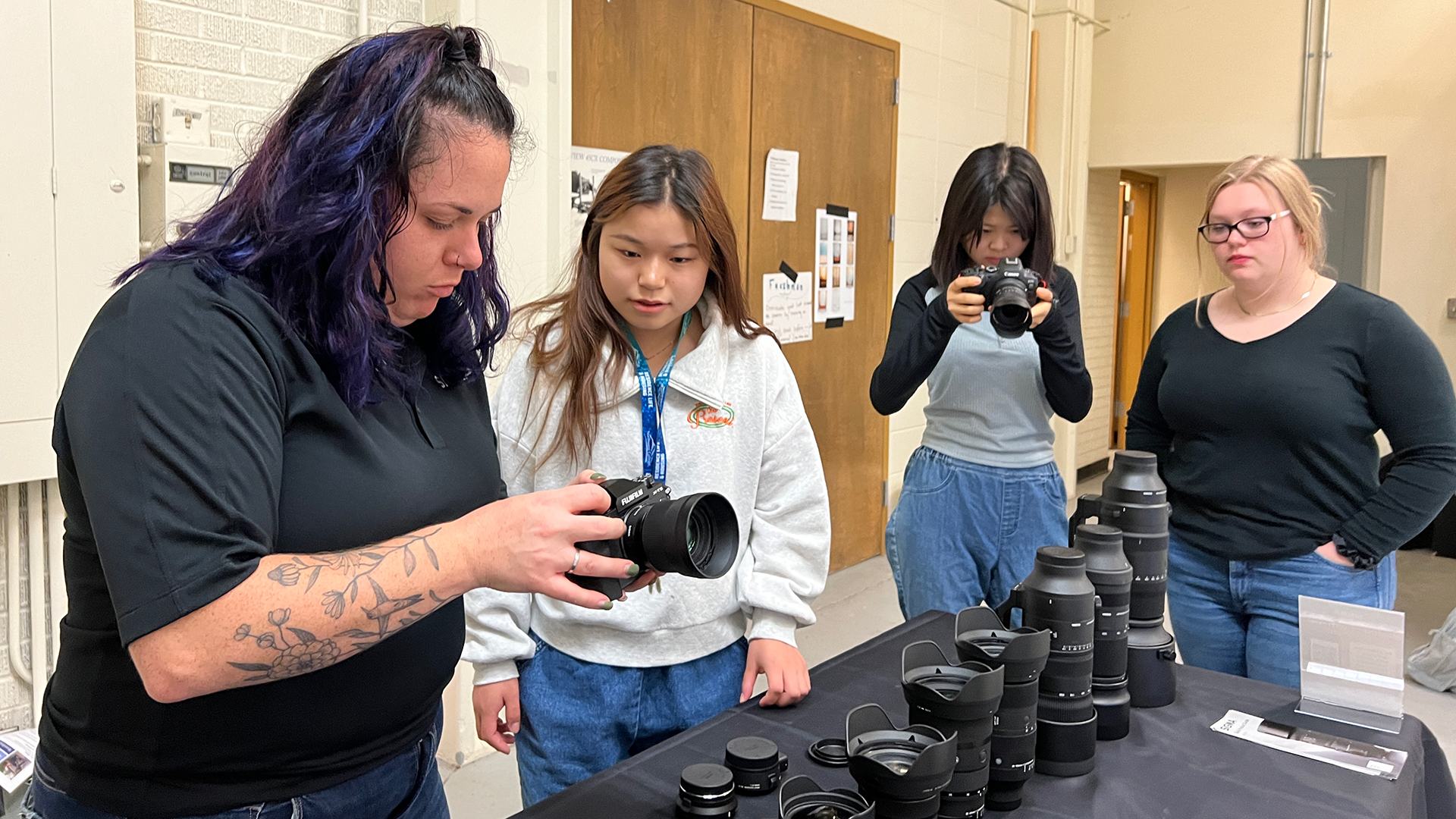A woman with dark hair in a dark shirt holding and looking at a camera, showing it to a female student while standing behind a table filled with other lenses, and two other students standing next to the table looking at camera equipment