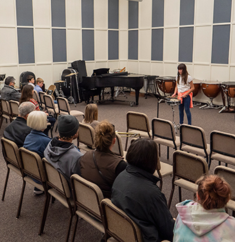 A group of people sitting in chairs in a classroom listening to a young female student playing drums with other instruments behind her