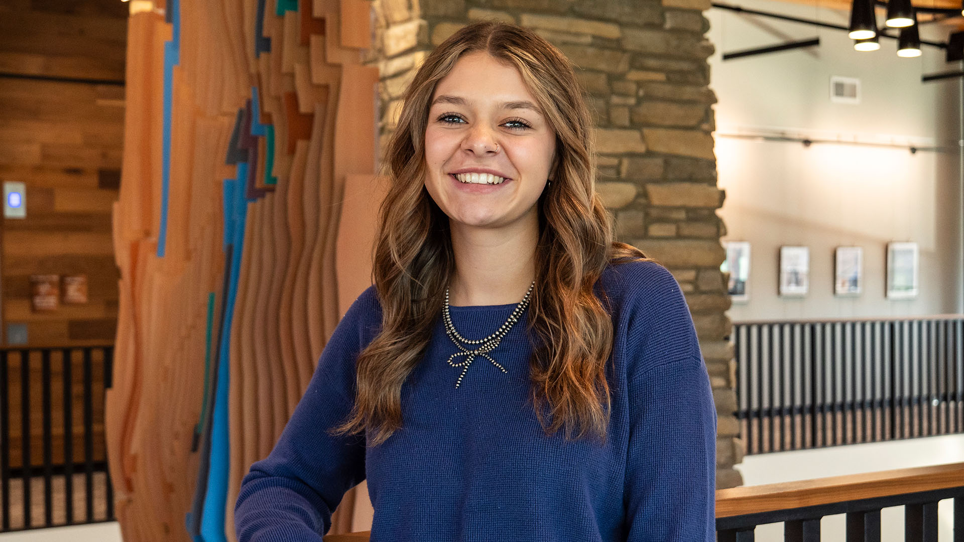 A smiling female student wearing a blue sweater standing against a second floor railing