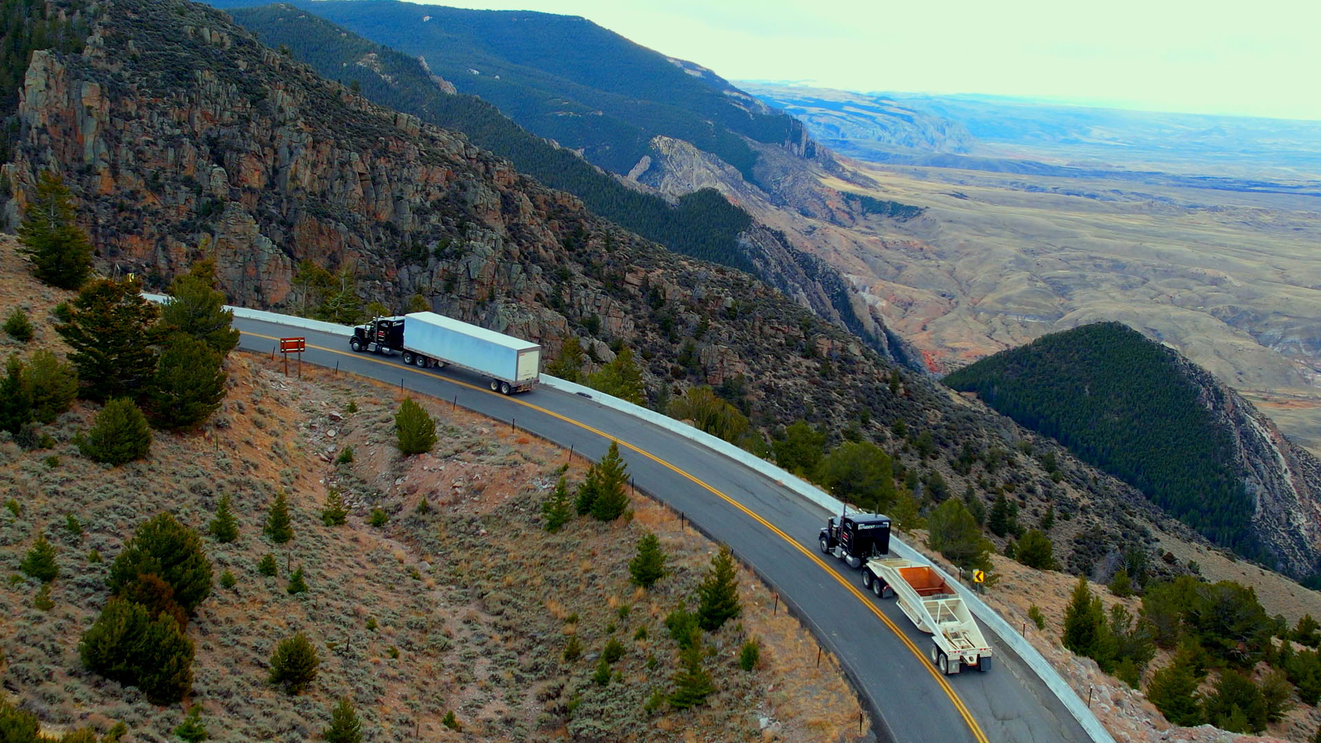 Two semi trucks, driving up a curving mountain road, seen from above