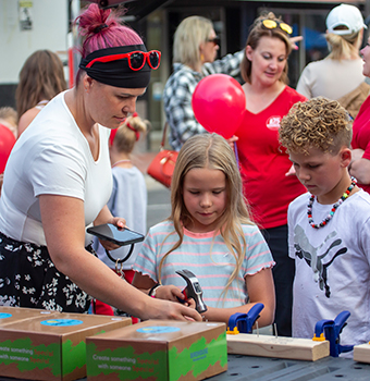 A young boy and girl being shown how to build something with a hammer and wood by a woman