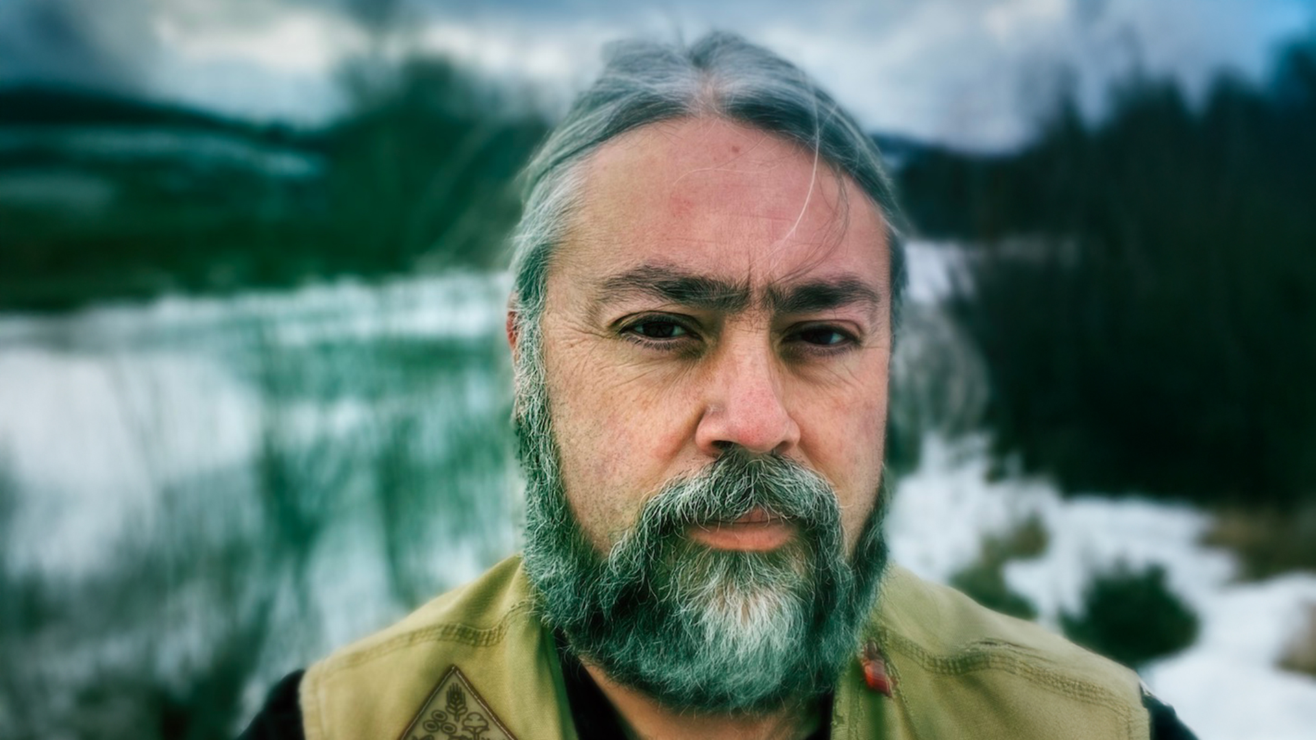 A man with a dark beard streaked with grey against the backdrop of a dark cloudy sky and snow covered landscape