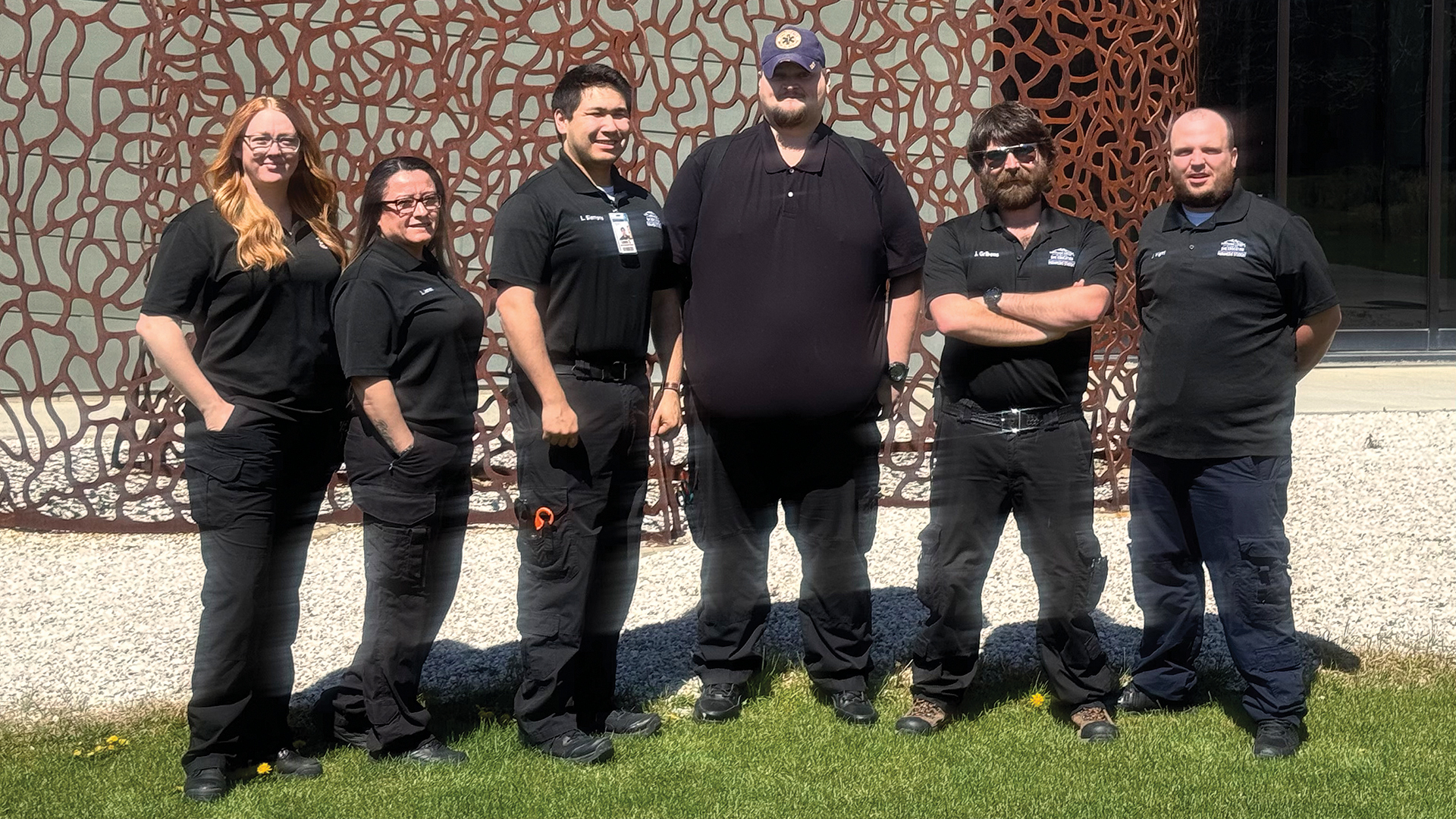 A group of two females on the left and four males on the right standing in front of a piece of lattice artwork in the grass