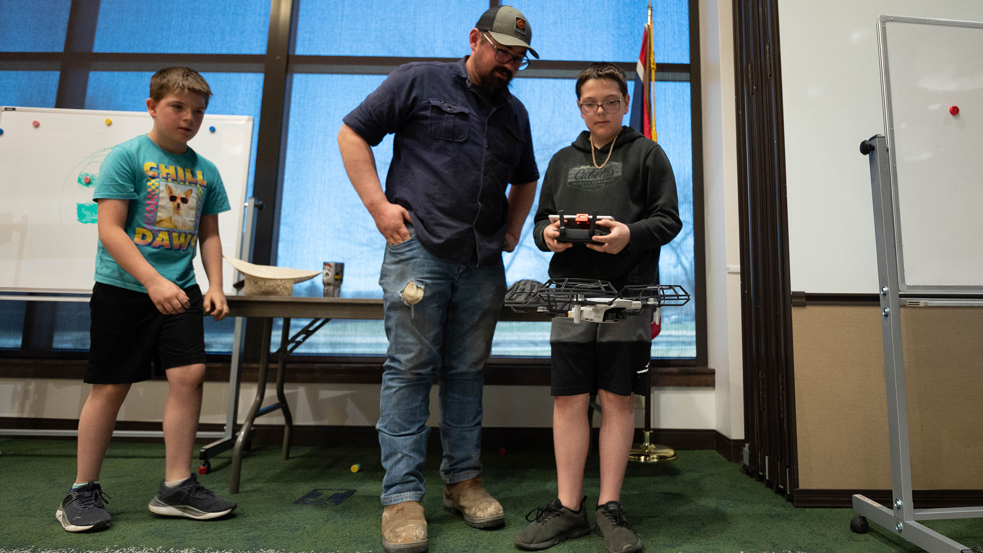 A man watching a boy control a small drone in a conference room with a second boy watching