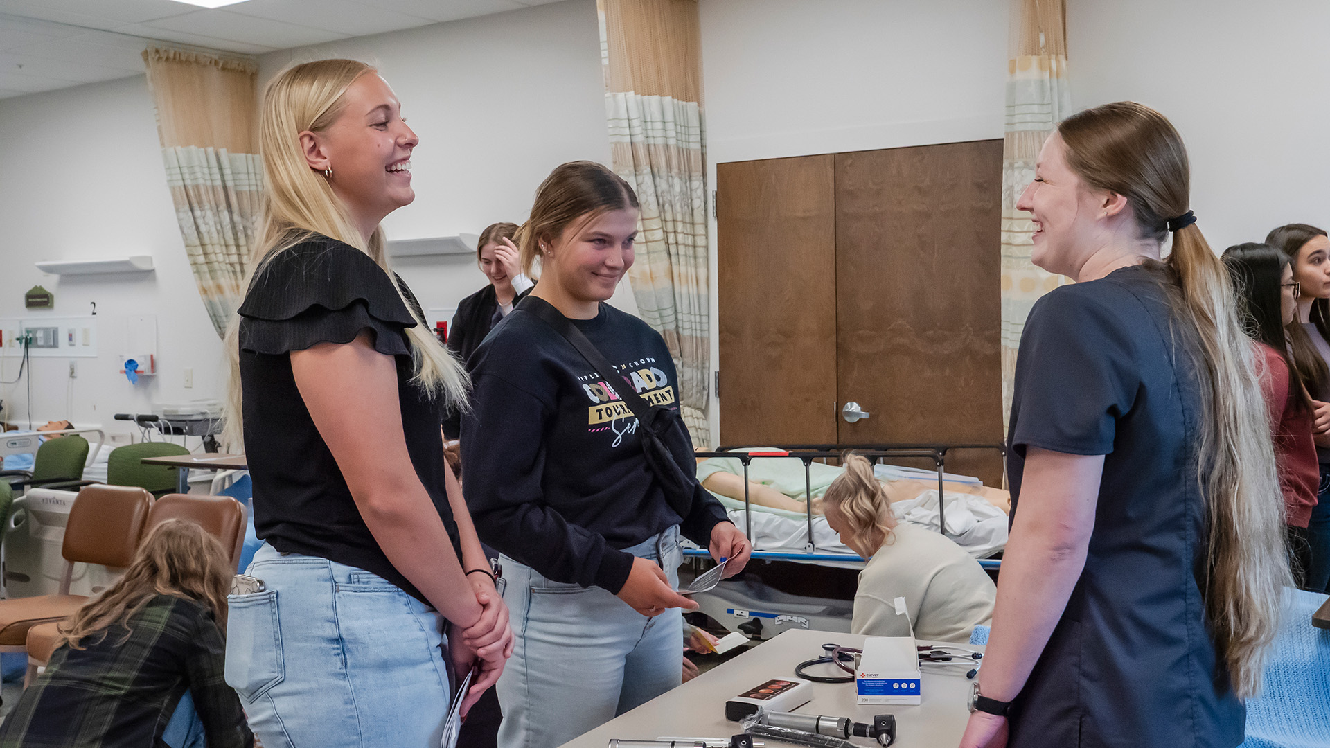 Two female high school students in a nursing lab smiling while talking to a female college nursing student