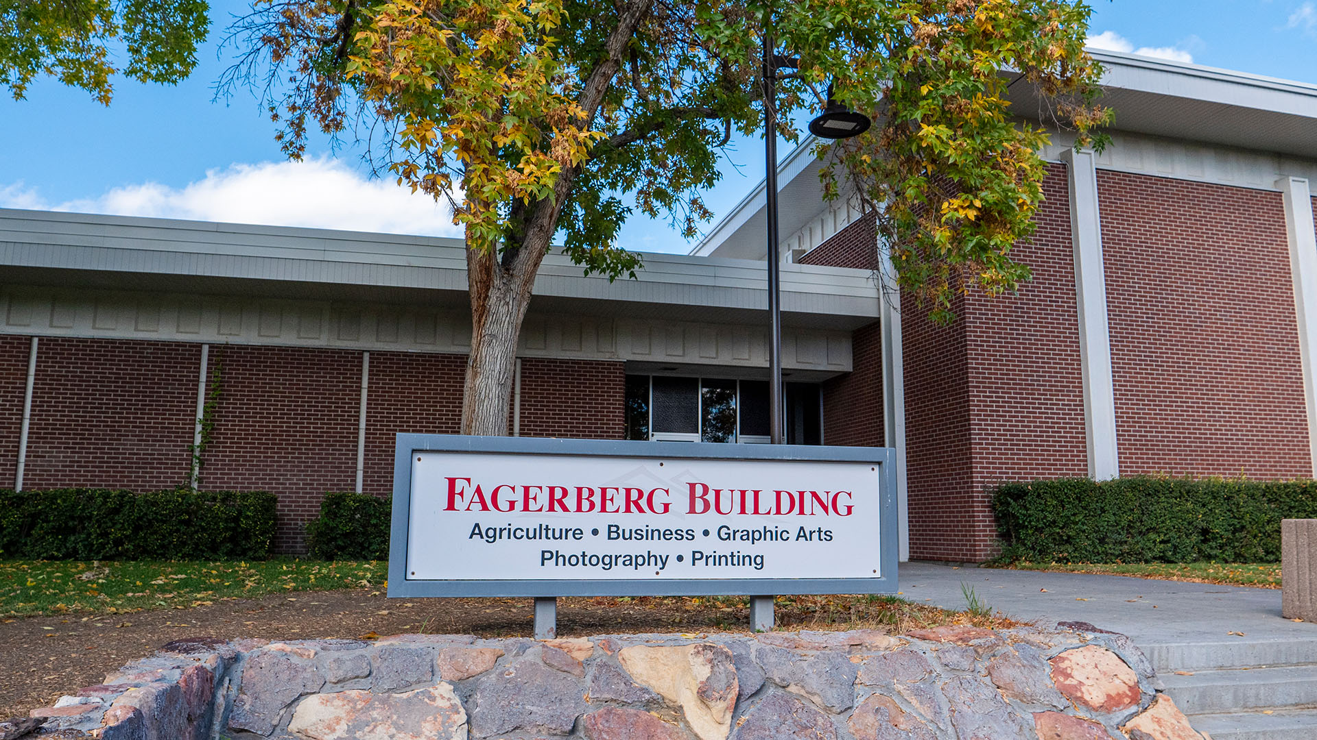 Outside the Fagerberg Building with a sign with red lettering on top of a stone shelf with red-bricked building, fall colors, and a blue sky in the background