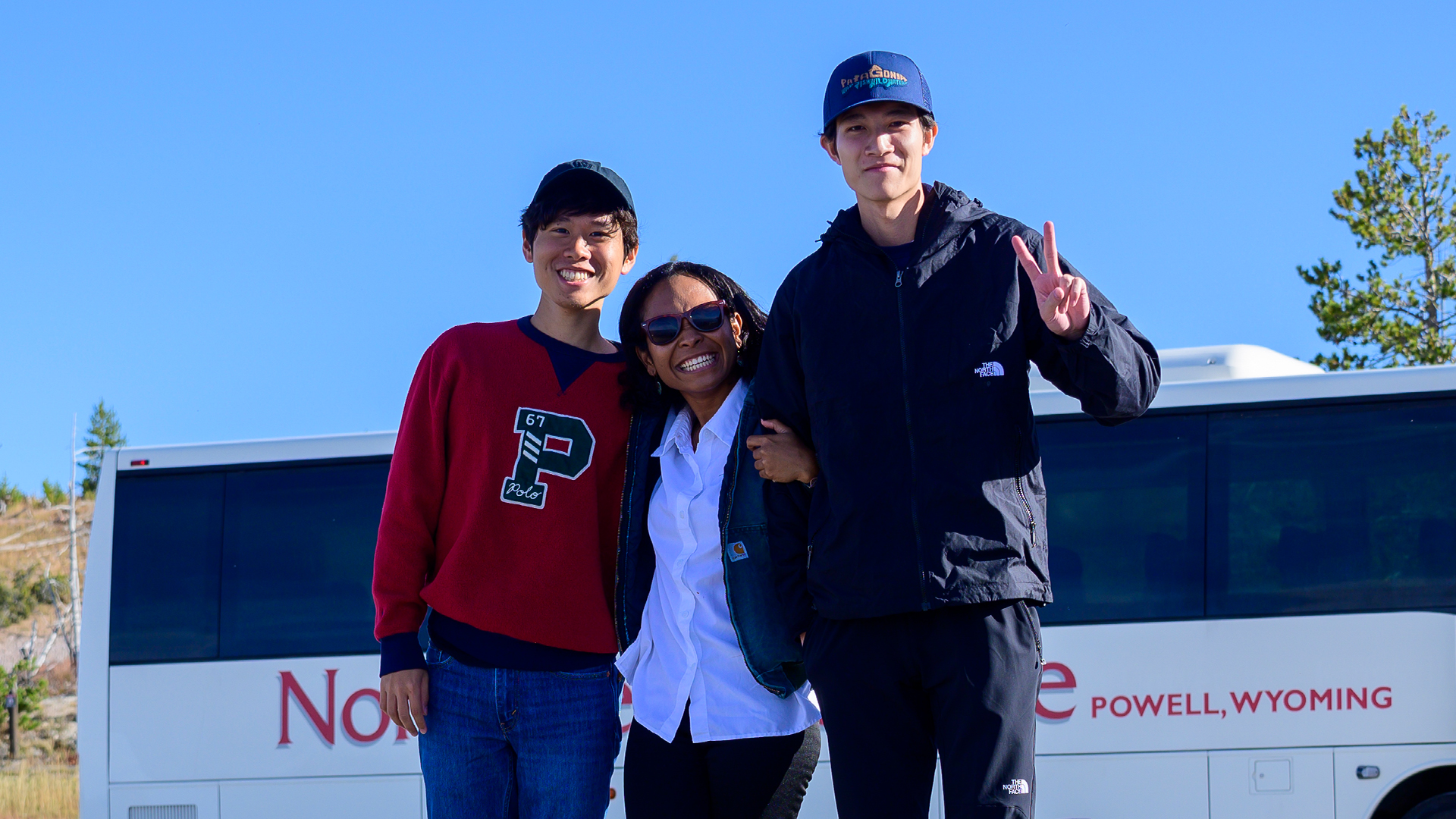 Three international students, a female in between two males who are wearing hats, with a white Northwest College bus and a blue sky in the background