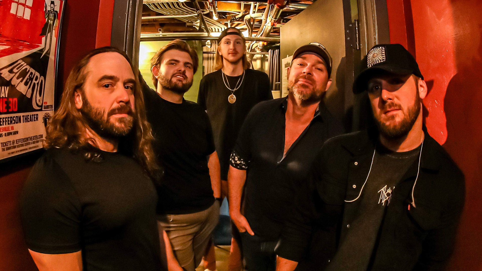 Five men, all with beards, some with baseball hats standing in a hallway looking at the camera, with two on the left, two on the right and one behind them in the center