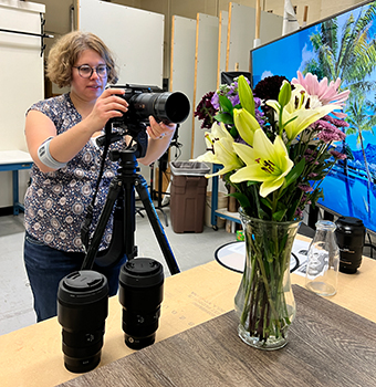 A woman wearing glasses standing behind and holding a camera on tripod pointed at a vase of flowers on a table with two other lenses to the side of the vase.