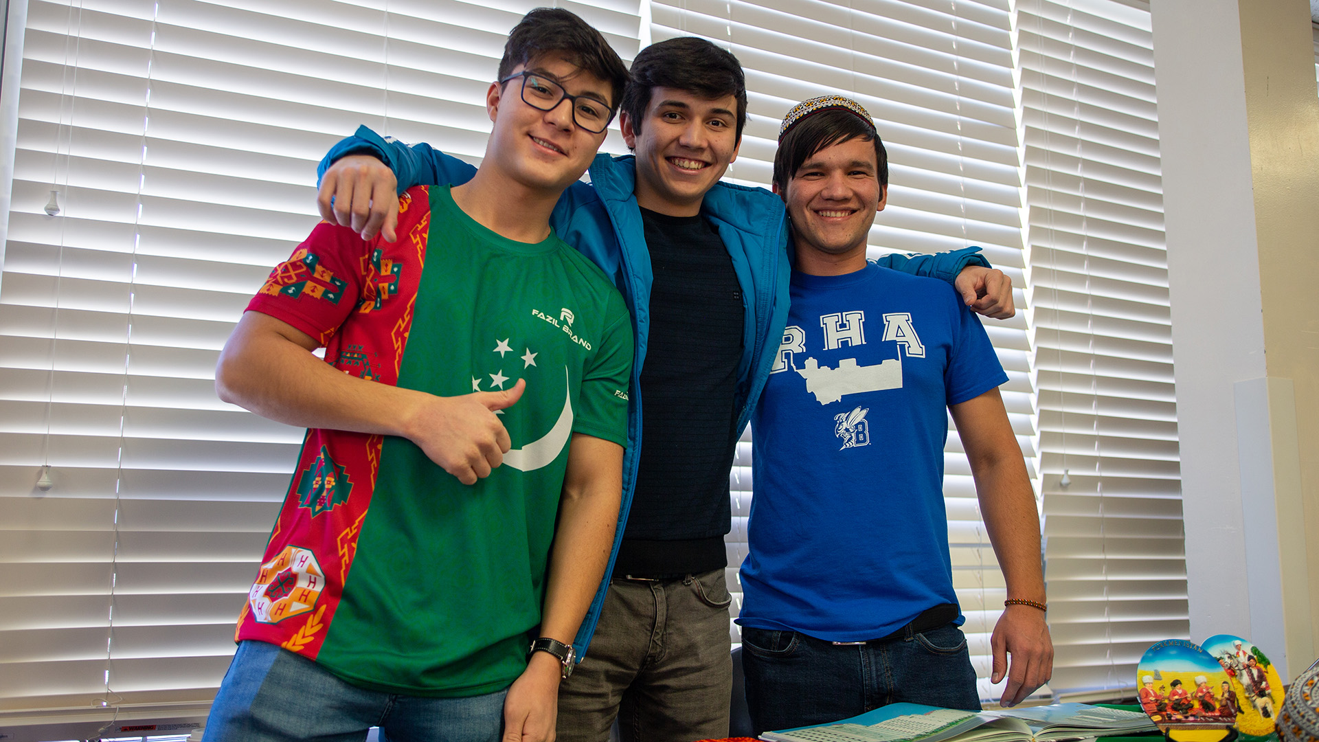 Three male college students with arms around each other's shoulders, standing behind a table with art in front of blinds