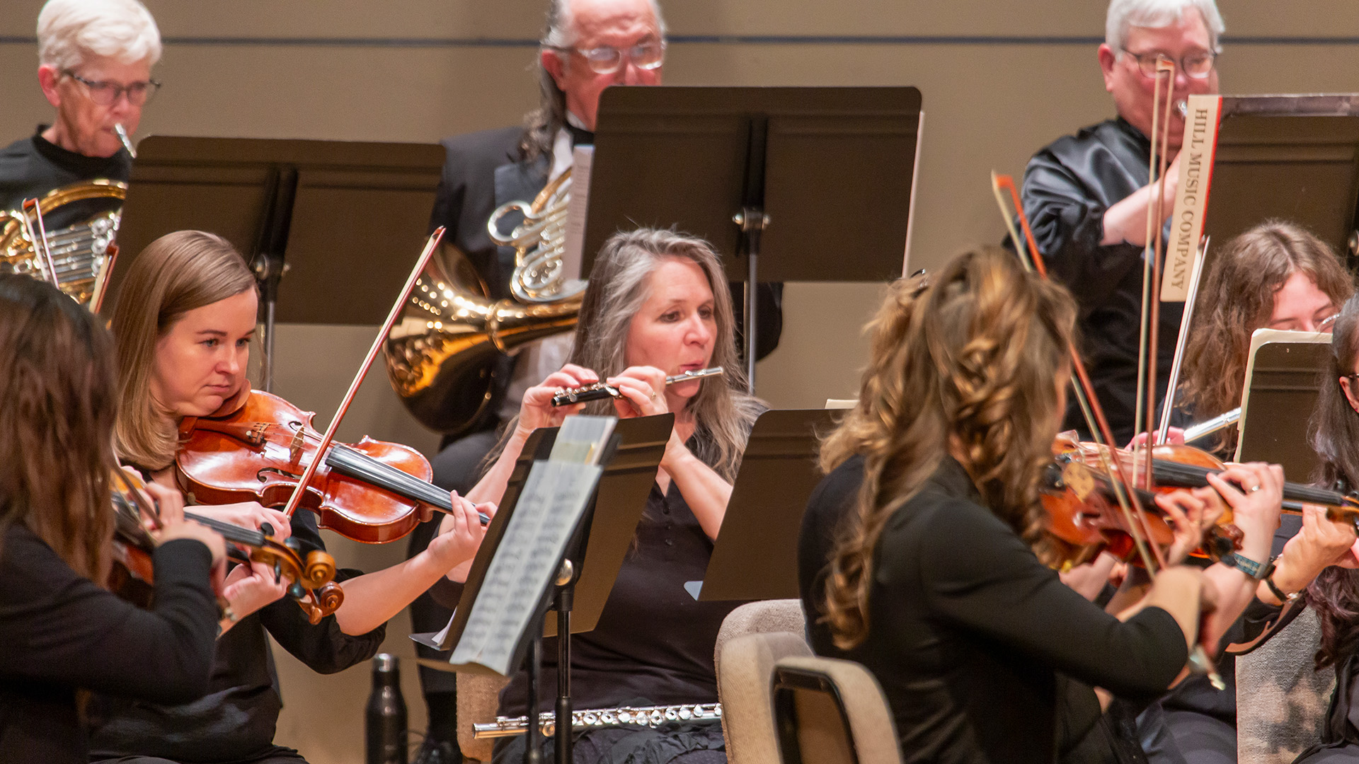 A woman playing a flute surrounded by other orchestra members with violins and brass instruments