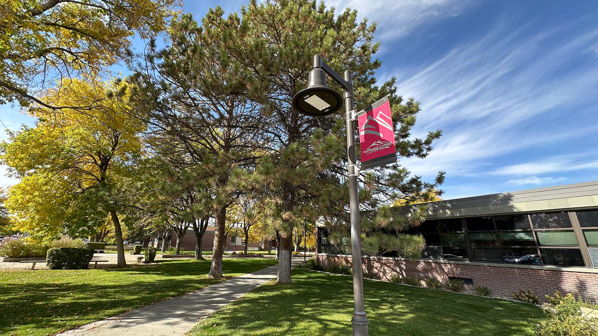 Northwest College banner on a light pole against a blue sky with wispy clouds along with pine trees on a fall afternoon