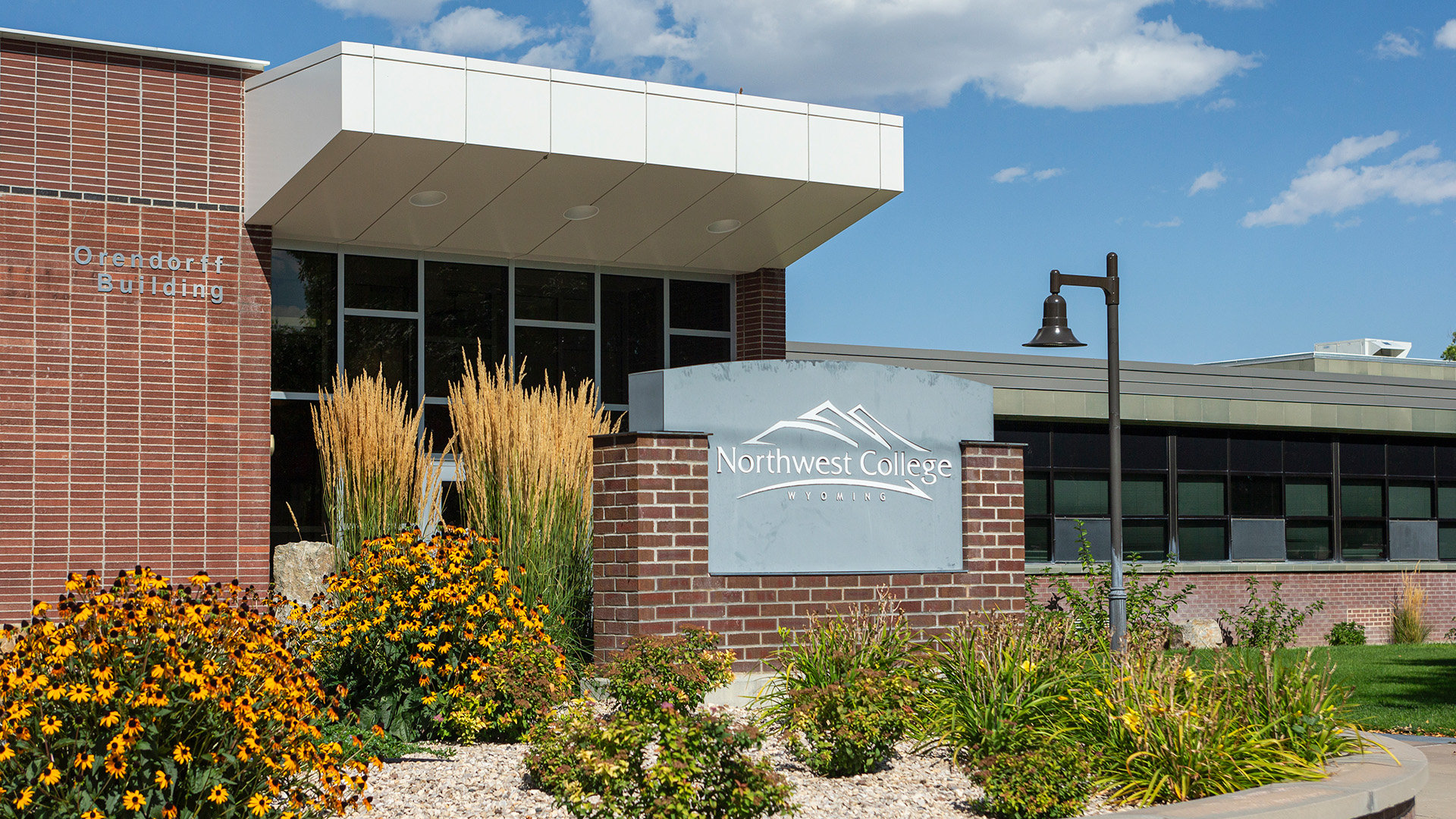 Outside front entrance of the Orendorff Building with grey Northwest College sign inside a small brick wall surrounded by rocks and flowers