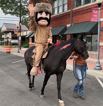 Trapper mascot riding a brown horse waving to the crowd