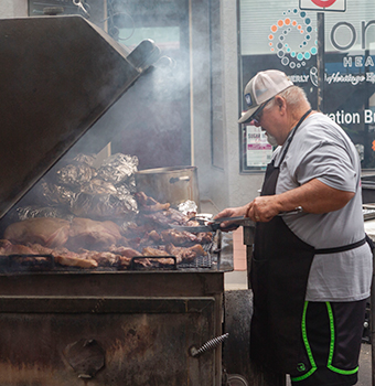 A man in a baseball hat and apron grilling different meats with smoke rising from the large open grill
