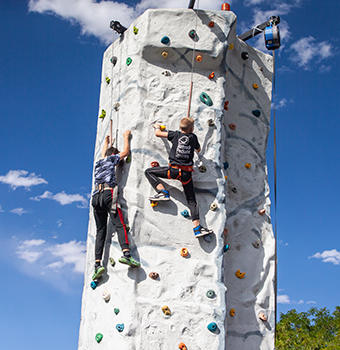 Children near the top of a climbing wall against a partly cloudy blue sky
