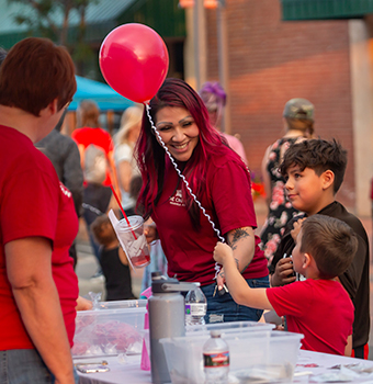 A child receiving a balloon from a woman at a table with another smiling child watching